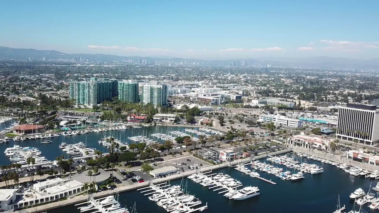 Marina del Rey port with luxury yachts and boats during sunny day. Los Angeles downtown in background. Luxury condos and residence apartments near busy Main Street. Aerial wide shot