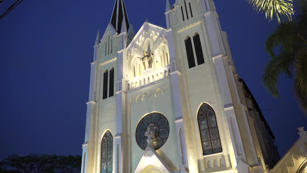 Panorâmica ascendente da fachada iluminada de uma igreja neogótica ao anoitecer, em um cenário urbano de Malang.