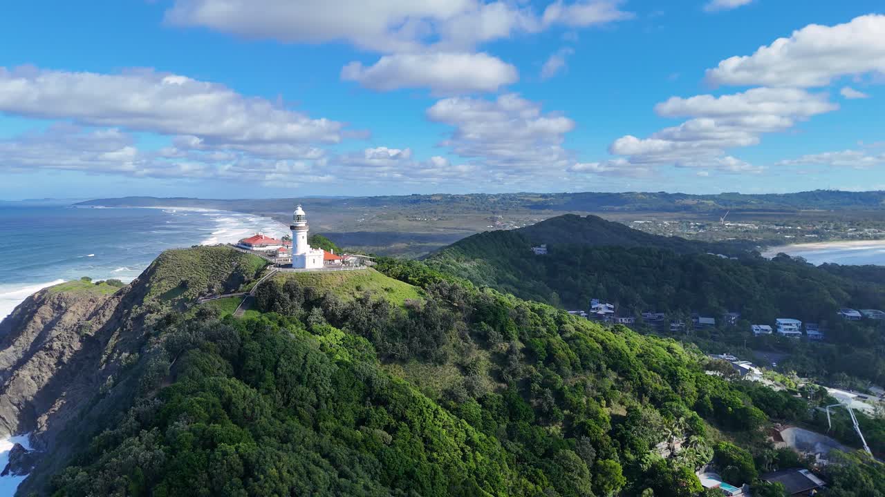 Drone footage captures Byron Bay lighthouse atop lush cliffs with ocean views under a bright blue sky
