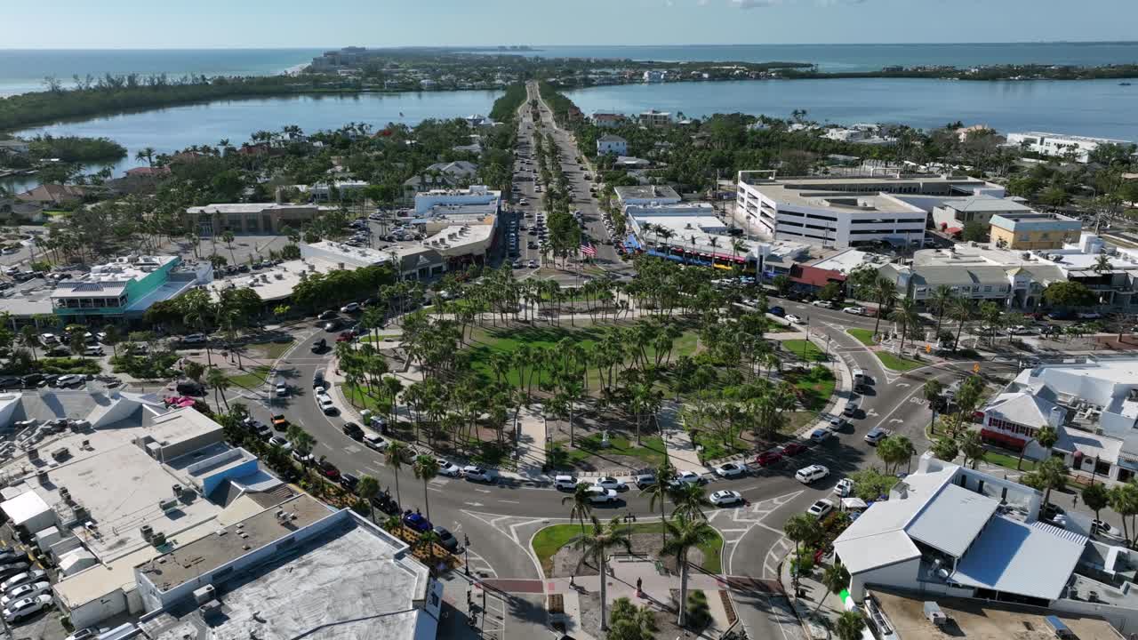 Aerial view of St. Armands Circle in Sarasota, showing vibrant shops, lush greenery and nearby waters. Bustling roundabout is surrounded by scenic landscapes under a clear blue sky. Sarasota, Florida.