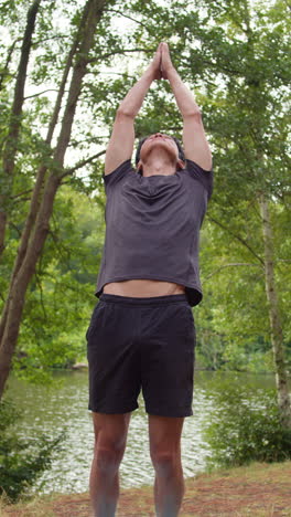 video vertical de un hombre con ropa deportiva haciendo yoga se estira en una alfombra en el bosque junto a un lago o río disfrutando de la paz y la belleza de la naturaleza filmado en tiempo real 2