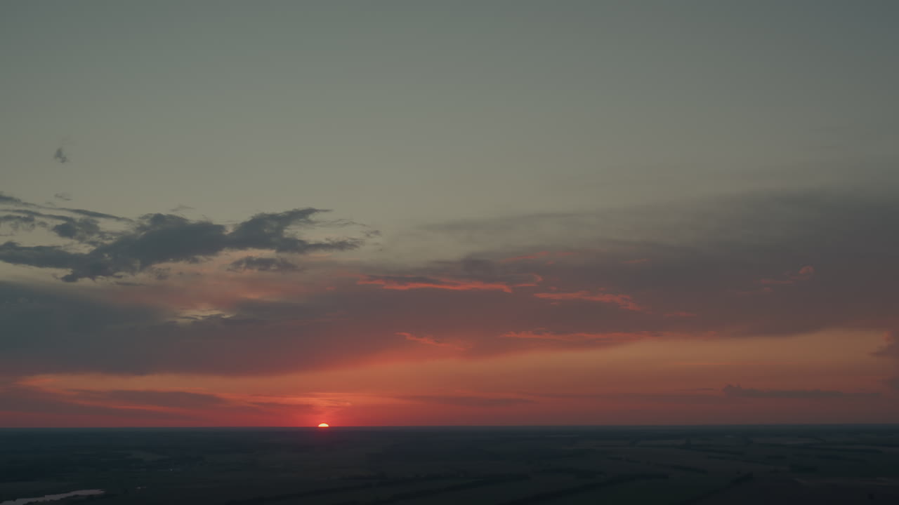 Vibrant orange red sky with dark dusk clouds framing half setting sun over expansive flat fields horizon with subtle lens flare and serene evening atmosphere, tinted pastel glow and calm mood