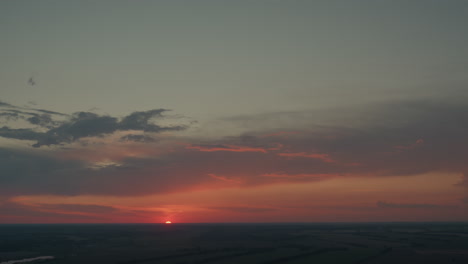 vibrant orange red sky with dark dusk clouds framing half setting sun over expansive flat fields horizon with subtle lens flare and serene evening atmosphere, tinted pastel glow and calm mood
