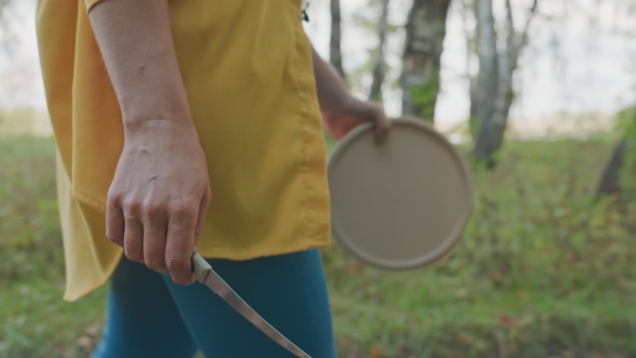 White woman holding knife camping pan, closeup of hand gripping serrated blade beside lightweight pan at campsite, focused camp cook preparing meal with steady motion, rustic forest edge and grass