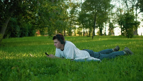 side view of young man lying on grassy field wearing headphones, engrossed in his phone, nodding his head as he enjoys music in a peaceful park surrounded by lush green trees