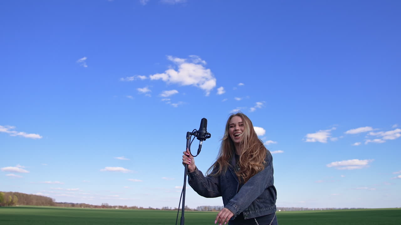 Portrait of a happy female singer on field. Young beautiful woman in denim suit with long hair standing next to the microphone on blue sky background.