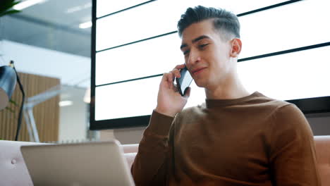 Young-businessman-using-phone-working-on-a-laptop-computer-sitting-on-a-sofa-in-a-casual-office,-close-up,-low-angle