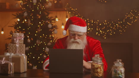 Real-Santa-Claus-using-new-technology-for-communication-with-children-receiving-mail-or-wish-list.-Cheerful-working-on-laptop-and-smiling-while-sitting-at-his-chair-with-fireplace-and-Christmas-Tree.-High-quality-4k-footage