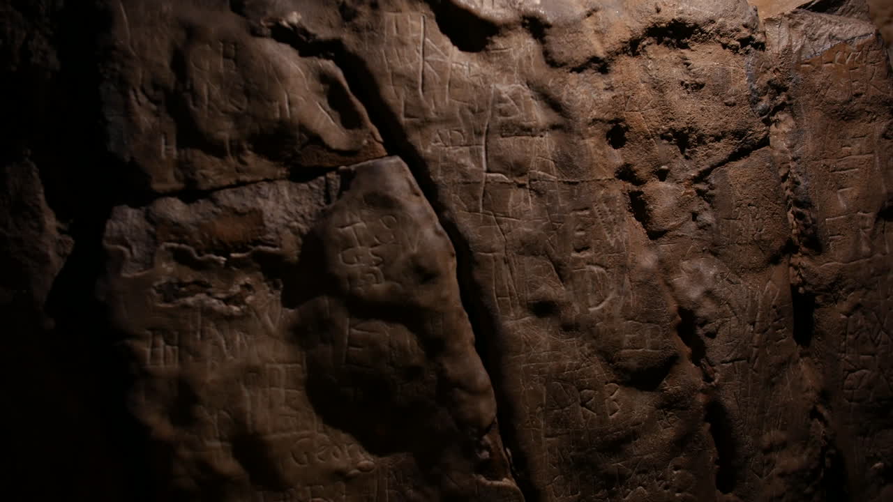 A cave wall with carvings, runes and inscriptions etched into the stone. The uneven surface and dim lighting add to the ancient and mysterious atmosphere. Marks left by past visitors and explorers