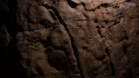 a cave wall with carvings, runes and inscriptions etched into the stone. the uneven surface and dim lighting add to the ancient and mysterious atmosphere. marks left by past visitors and explorers