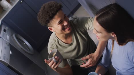 happy biracial couple sitting on floor in kitchen, drinking coffee