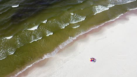 two people relaxing under rainbow colored umbrella lying on white and fine sand with waves gently rolling onto the shore of baltic sea