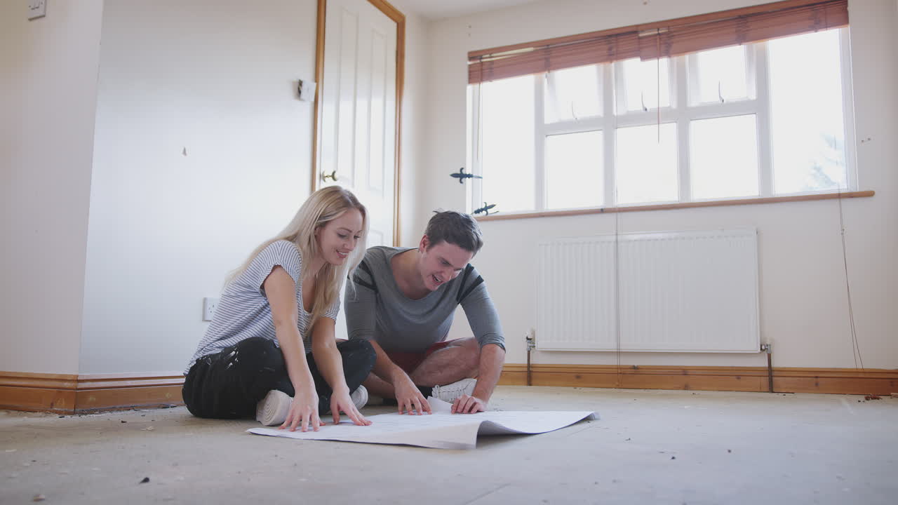 Pareja sentada en el suelo mirando planos de planta en una habitación vacía de su nuevo hogar.