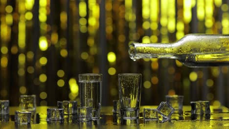 barman pour frozen vodka from bottle into shot glass. ice cubes against shiny gold party background