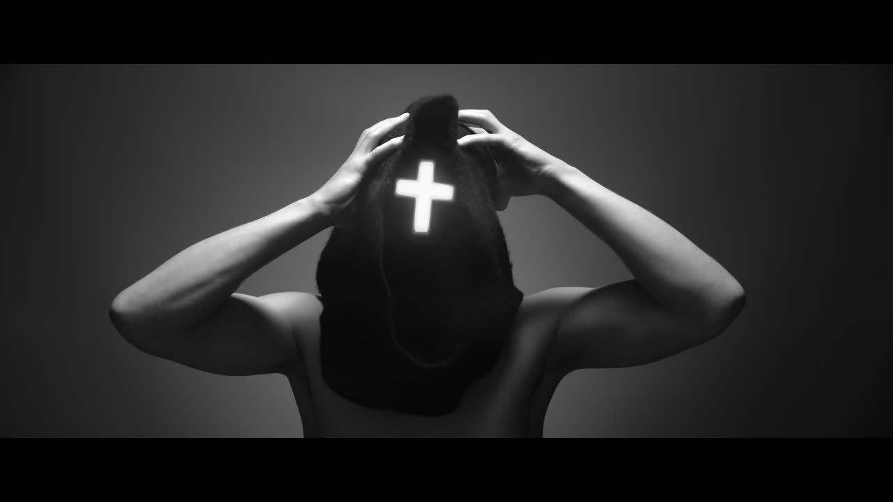 Masked man with cross of light glowing on his face grabs his head with his hand