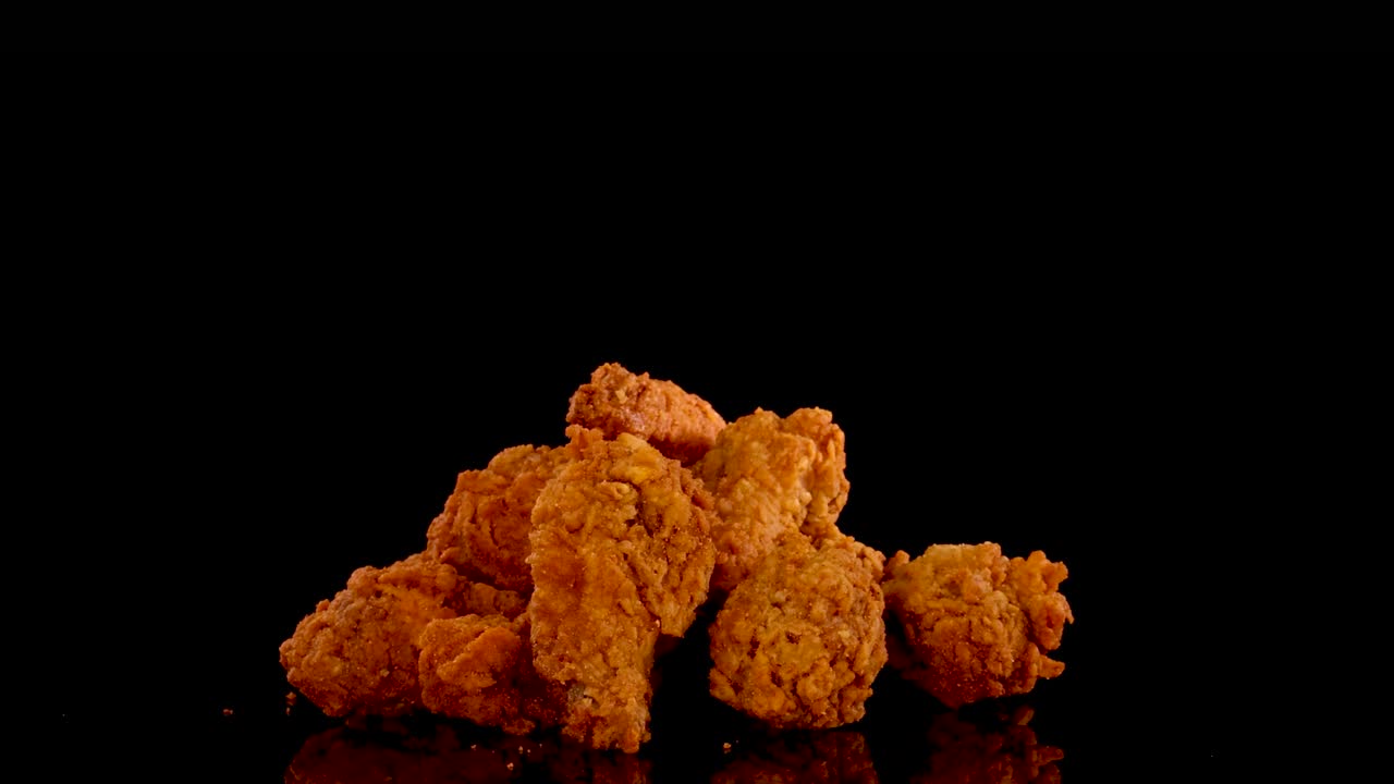 Golden, crispy fried chicken pieces are shown in a slow motion zoom, with dramatic lighting and a black background emphasizing texture and color