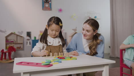 Little-Girl-In-A-Montessori-School-Playing-With-Shapes-Stacking-While-Teacher-Helping-Her