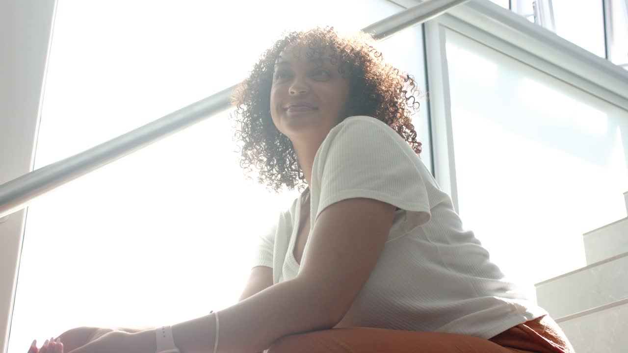 Donna afroamericana seduta sulle scale accanto al parapetto della finestra, abbassa un oggetto e sorride a una voce fuori campo.