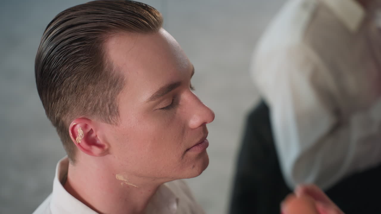 celebrity wearing foundation on neck and ear adjusts collar while makeup artist applies cosmetic product creating polished on-camera look with soft studio background