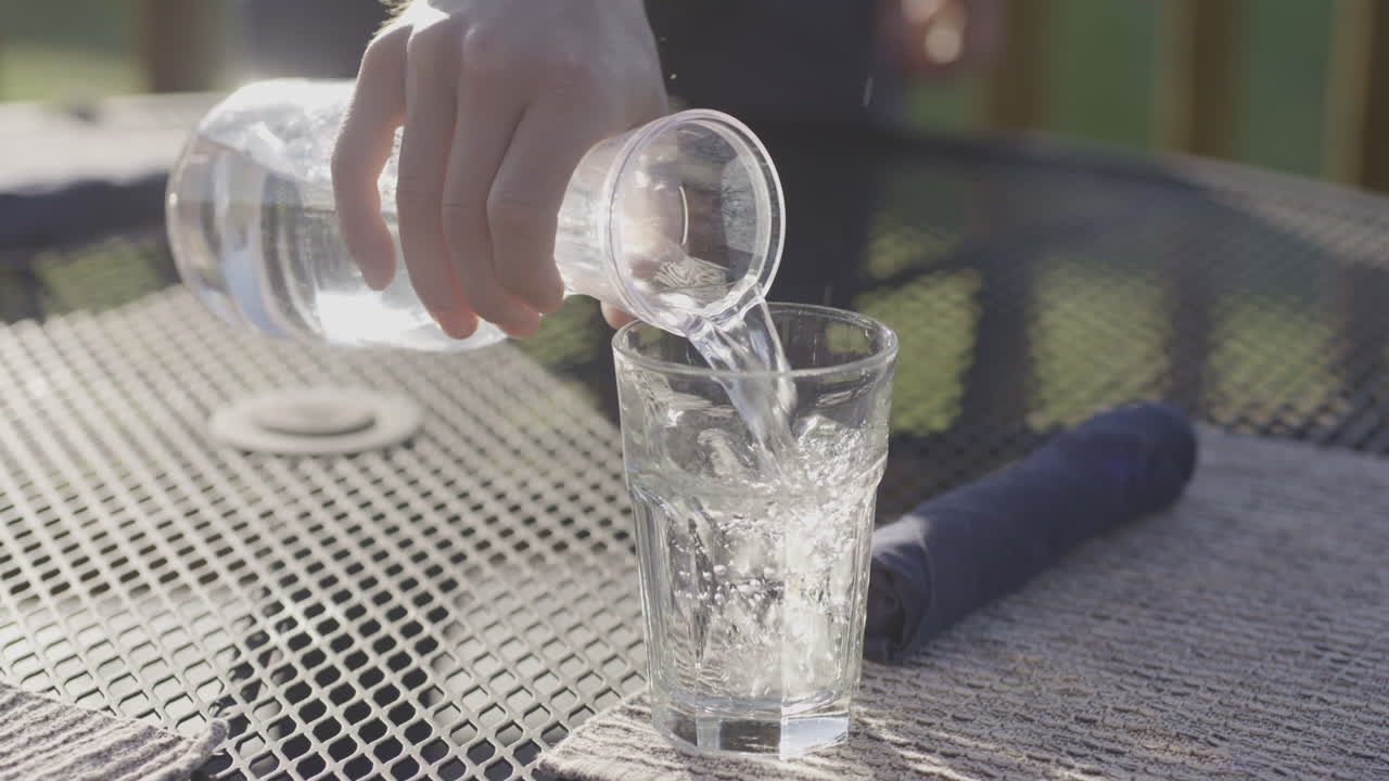 mano vertiendo agua con cubitos de hielo de la jarra al vaso en la mesa al aire libre