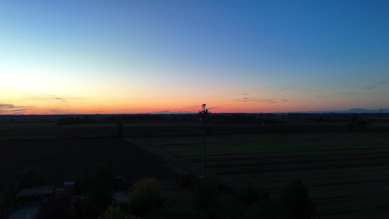 Un'antenna per telecomunicazioni si staglia contro un caldo tramonto su campi agricoli nell'Italia settentrionale, ripresa durante un'orbita di drone.