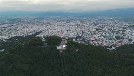 Seguimiento-Aéreo-De-Una-Casa-En-La-Cima-De-Una-Montaña-En-Primer-Plano,-La-Ciudad-Capital-De-Salta-Al-Fondo