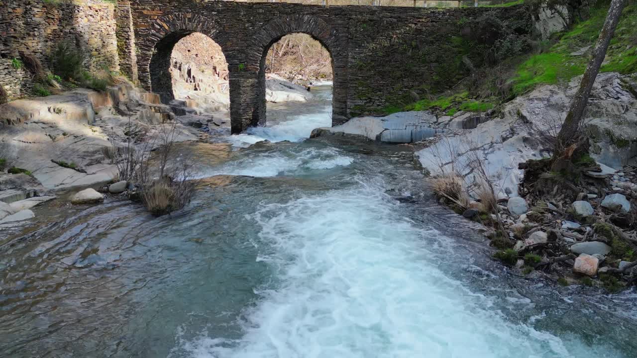 Imágenes aéreas de un río claro fluyendo bajo un puente de piedra histórico en Extremadura, España, con lecho rocoso y entorno natural. Paisaje escénico de viaje y patrimonio europeo.
