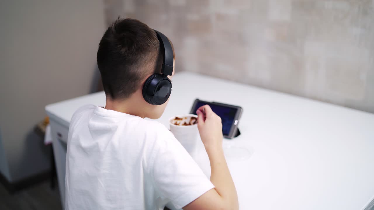 Boy watching video and eating at home. Back view of a child with headphones sitting at the table in the kitchen and looking at tablet.
