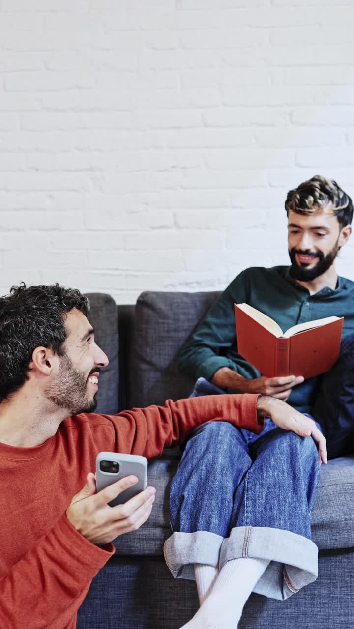 Dos hombres relajándose en casa, uno usando el teléfono y el otro leyendo.