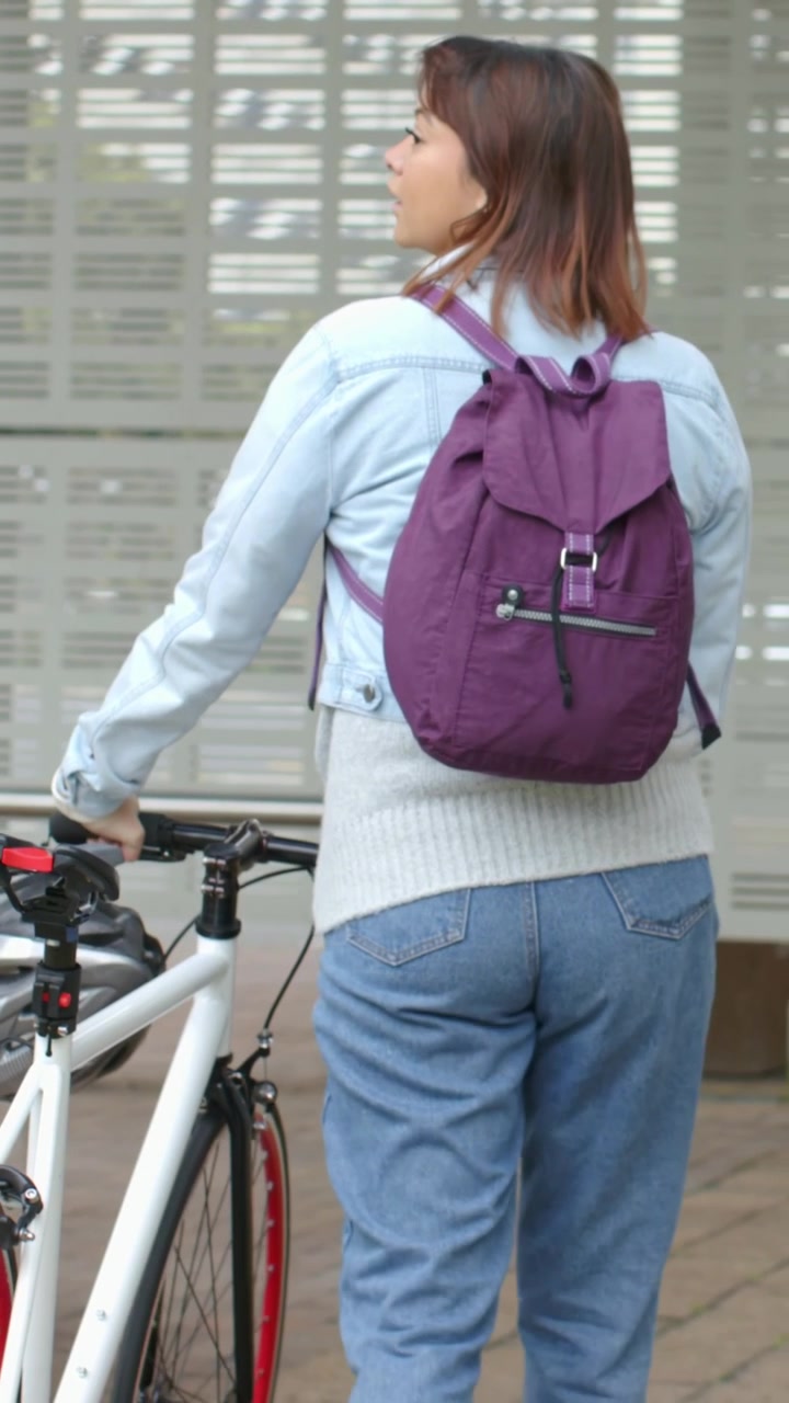 Vertical video: Walking woman pushing bike on path, denim jacket and purple backpack, checking road