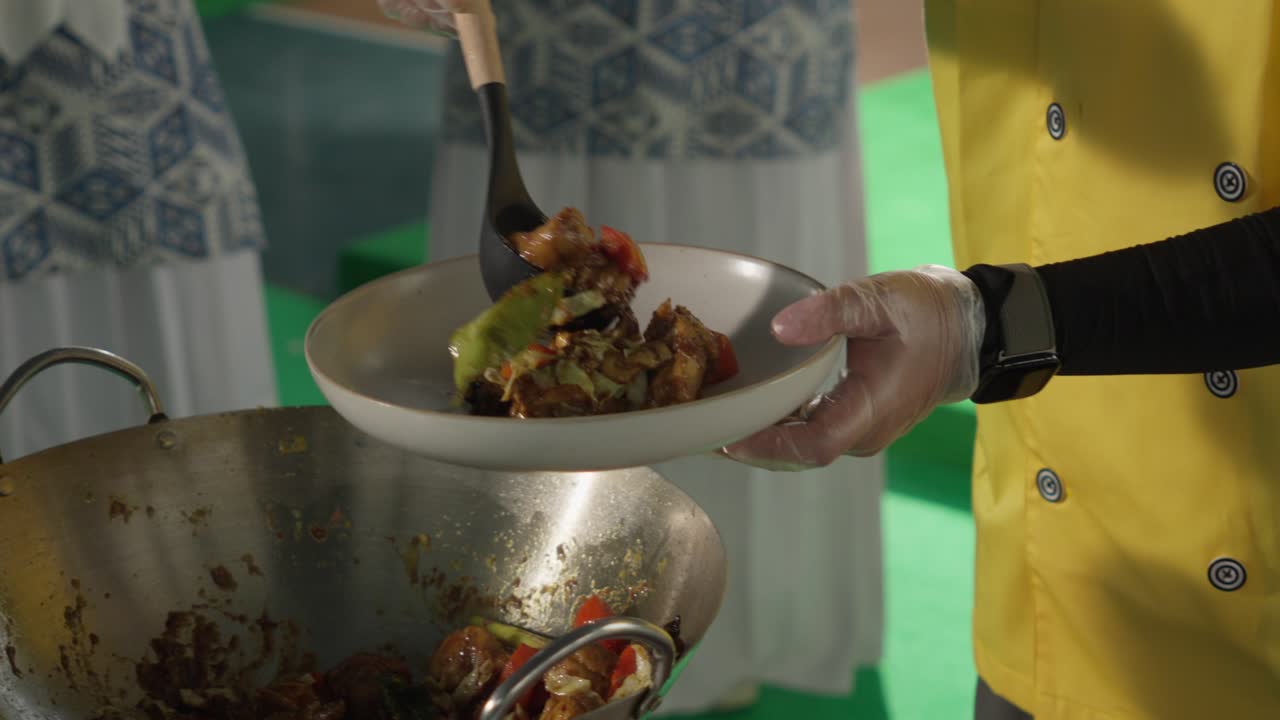 Chef In Yellow Uniform Serves Stir-fried Vegetables And Meat From Wok Into Plate Using Black Ladle. closeup shot