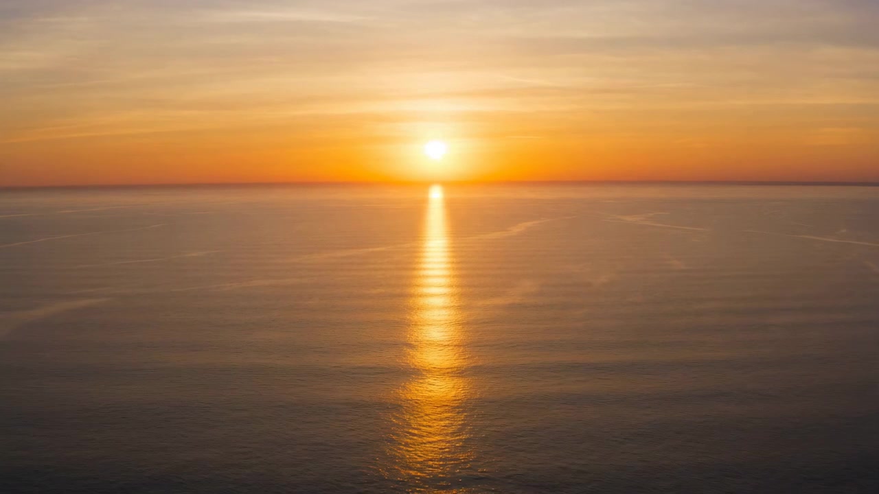 Sun sits centered above distant horizon, casting long golden reflection across smooth ocean beneath thin layered clouds and warm gradient sky, creating serene minimal seascape atmosphere with quiet