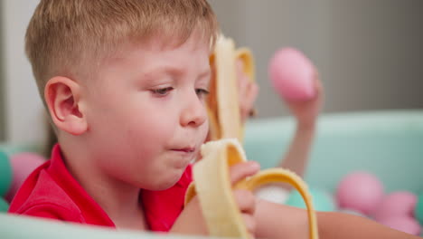 close up of young boy eating banana while sitting in pastel ball pit, playful moment captured with blurred view of girl nearby also enjoying banana and colorful pastel balls