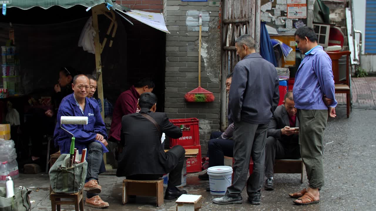 Five men having a work break in the middle of the day to eat, rest and talk in a back alley with a woman in the background, handheld shot