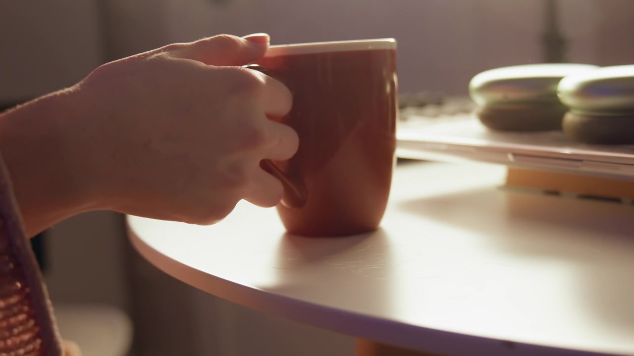 Two hands lift glazed ceramic mug from round tabletop while warm side sunlight creates soft highlights, gentle shadows, blurred dishes, and relaxed home mood with clear copy space, with hand, mug
