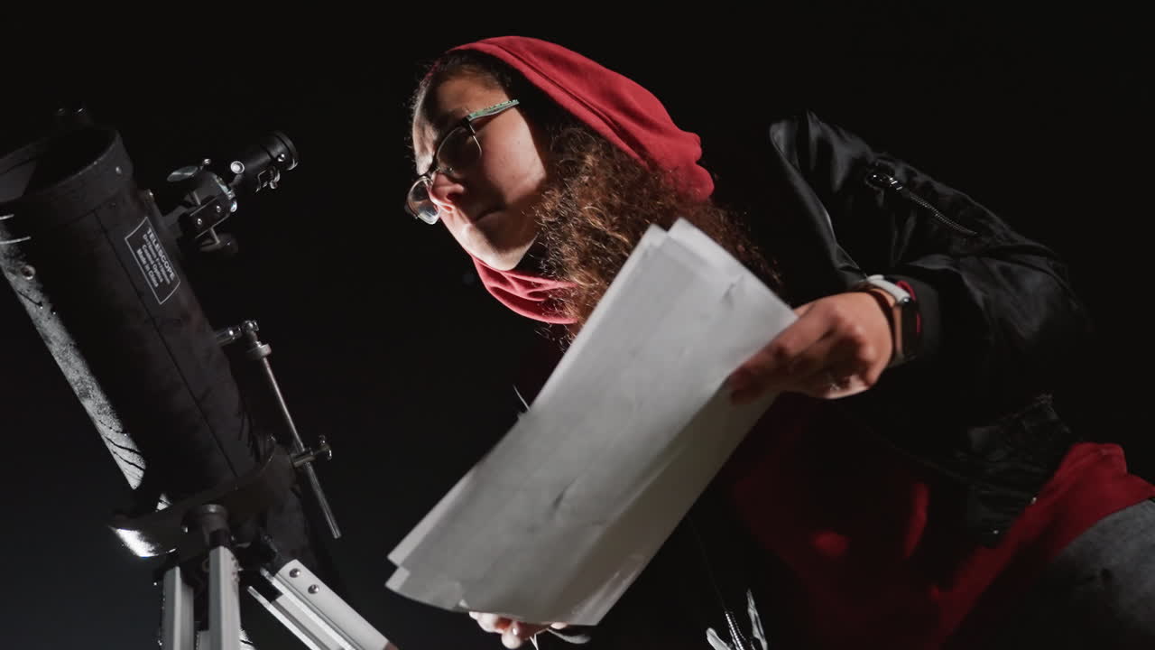 night stargazer checks paper star chart, leaning over telescope, red hoodie and glasses visible, hands flipping printed maps, tripod and lens in frame, focused expression, quiet atmosphere