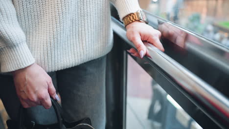 lower angle view of mindful explorer in knit sweater and jeans wearing gold chain watch and holding handbag while descending glass elevator, hands on railing reflecting modern store interior floor