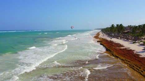 Drohne-Fliegt-über-Türkisblauen-Wasserstrand-In-Mexiko-Mit-Touristen,-Die-Schwimmen-Und-Paragliden