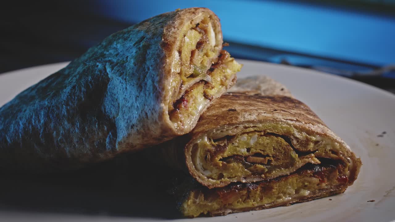una tortilla enrollada y rellena con pimiento morrón salteado, huevo y cebolla, cortada en dos mitades y apilada en un plato blanco con luces que iluminan el lado derecho del plato