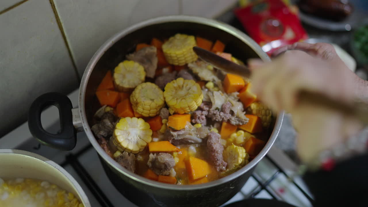 Colorful vegetables float in thick stew as Argentinian locro simmers in black pot, top shot overview of grandma hands