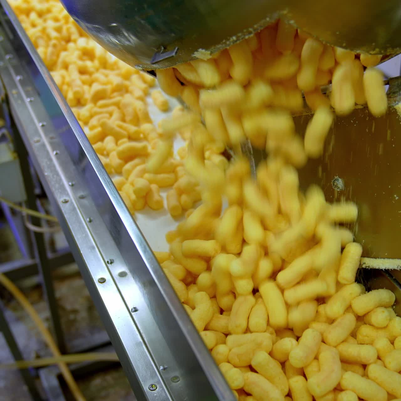 Lots of yellow crispy corn sticks fall on the conveyor line. Factory worker pushes snacks along the belt. Food factory production concept