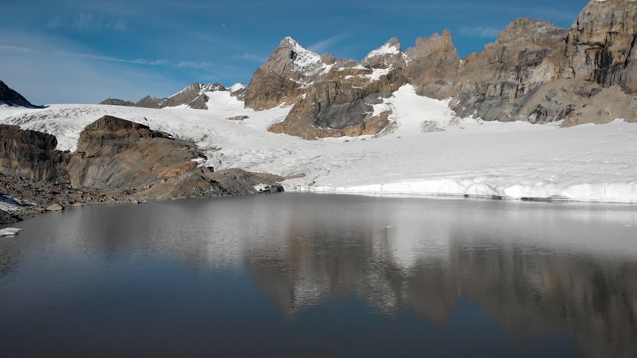 Aerial flyover over the glacial lake next to Claridenfirn glacier in Uri, Swizerland on a calm, peaceful morning with alpine peaks behind a wall of ice and icebergs floating in the water