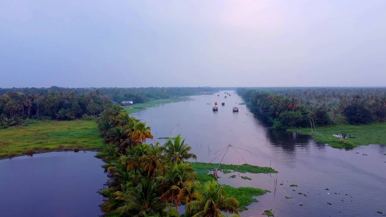 Bredt luftfoto af en snoet Kerala-kanal omgivet af tætte kokospalmer og grønne planter, der viser adskillige husbåde, der sejler langs den fredfyldte vandvej