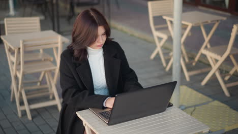 야외 쇼핑몰에서 노트북으로 일하는 전문 여성, 초점을 맞추고 세련된 검은 코트를 입고, 나무 의자와 테이블이 배경에 끔하게 배열되어 있습니다.