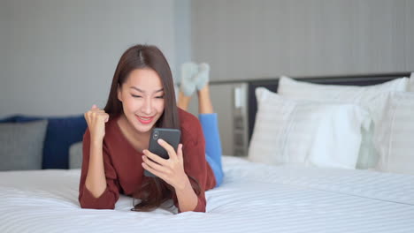 스마트폰을 손에 들고 침대에 누워 웃고 있는 젊은 아시아 여성