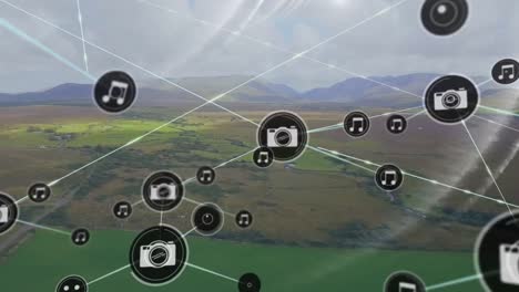 Animation-of-icons-connected-with-lines,-aerial-view-of-green-field-against-mountains-and-cloudy-sky