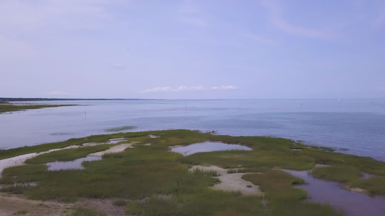 The camera pans from left to right in front of the dune grass, and tidal pools. A kayaker can be see out by the channel markers in the bay.