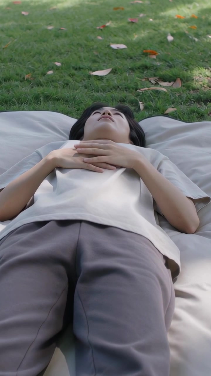 Video verticale: Luce che cambia, donna su un tappetino nel parco che inclina la testa e guarda gli alberi, con un top beige.