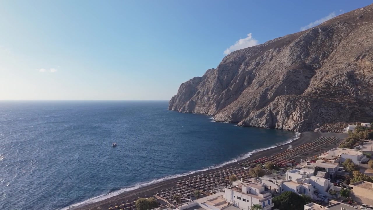 Filmato aereo laterale con drone della Spiaggia di Kamari a Santorini, che cattura la costa, la spiaggia sabbiosa, le scogliere e il blu profondo del Mar Egeo, lungo il paesaggio vulcanico dell'isola.