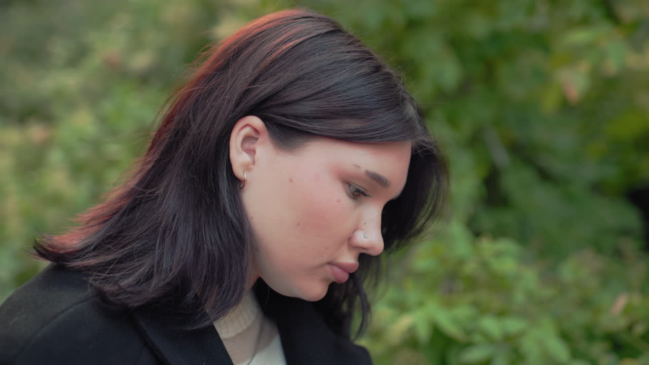 자연을 사랑하는 사람의 중간 측면 시점, 책 읽기에 집중, 공원 벤치에 앉아 머리카락을 귀 옆으로 정리, 부드럽게 흐릿한 숲 배경, 조용한 마음 챙김과 학습 분위기